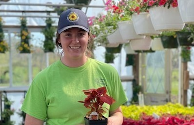woman with green shirt and blue hat standing in greenhouse holding plant